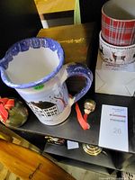Photo showing a white ceramic pitcher with a blue handle and rim, decorated with a snowman and winter houses, grouped with other holiday items including ramekins and a wooden box