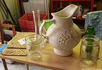 Photo showing white ceramic pitcher, two clear glass canning jars, tall glass vase, and woven coasters on table.