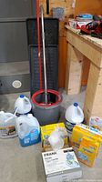 Photo showing mop bucket with mop and metal wringer, two black boot trays behind, and multiple containers of cleaning liquids gathered around the bucket on the floor.