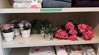 Wide view of nine faux flower arrangements on shelf including plastic and pottery pots and two bunches of flowers.