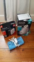 Photo of all three items: Noma oscillating heater in black box, Noma ultrasonic humidifier in white and light blue box, and a blue heating pad with attached white control switch cord laid on a wood floor.