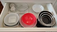 Top view of nested stainless steel mixing bowls, white ceramic bowls, clear glass bowl, red plastic bowl, ramekins, and plates in drawer