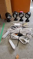 Four black Netbrite outdoor motion sensor lights with adjustable bases and several bundle white extension cords with plugs and switches on concrete floor.
