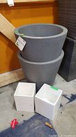 Two stacked gray concrete self-watering planters with two white square ceramic pots in front on the floor.