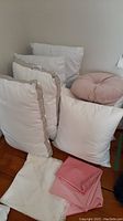 Lot showing the four white pillows and cushions with two pink fabric pillow covers and one embroidered white pillow cover on floor and chair.