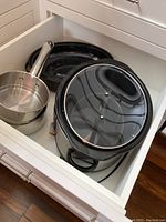 Draw filled with the black enameled roaster with glass lid, stainless steel saucepan and sauté pan, and additional cookware.
