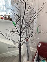 Artificial twig tree with multiple branches, dark brown color, dusted with fake snow, standing on a white cloth base with string lights.