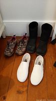 Top view of three pairs of shoes: white Vans slip-ons, Merrell hiking shoes, and black UGG boots on a wooden floor.
