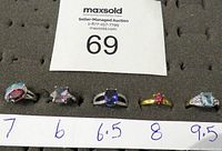 Five rings arranged in a row with size labels; stones in various colors including red, purple, blue, and aqua. Condition appears like new.