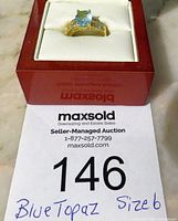 Blue topaz ring displayed inside a red jewelry box on white padding. Ring has a large oval blue topaz stone and multiple clear accent stones on the band.