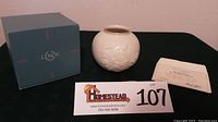 Photo showing original blue Lenox box, the round white vase with embossed floral design and certificate of authenticity on black cloth background.