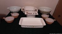Whole group of vintage mixing and baking bowls arranged on table: two large Pyrex mixing bowls in light blue, two Corning Ware Spice of Life pieces (rectangular baking dish and casserole), one Floral Bouquet rectangular dish, one round white baker, and four small Fire King Ware handled bowls in peachy beige.