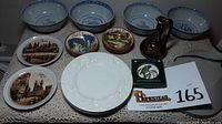 Lot displayed showing six blue and white Chinese soup bowls at back, two brown-toned Prague cityscape porcelain plates, two colorful signed Pueblo Español Barcelona bowls, white embossed ceramic plate, black and gold Greek art ceramic pitcher, and square coaster with bird design in original box.