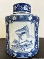 Full view of the ginger jar showing the main blue and white landscape design with tree, mountain, and building motifs, floral details on edges.