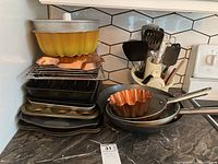 Overall view of assortment of baking and frying pans stacked together on countertop, showing yellow ceramic dish at top, copper mold, metal pans, and three frying pans with handles.