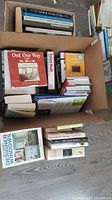 A box filled with multiple books, including visible titles like Out Our Way, Anne of Green Gables, and other assorted books in historical and self-help genres.