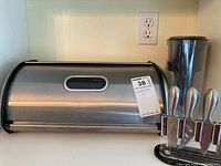Stainless steel metal bread box with a roll-top lid and viewing window, placed on counter next to oyster shuckers and Brita jug.