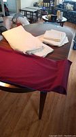 Solid wood dining table with burgundy table cover and white linens/place settings on top, showing detail of table edge and leg, wood grain visible.