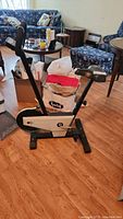 Stationary exercise bike positioned in a room with hardwood floor, chairs, and other household items visible in background.