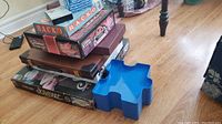 Stack of various boxed board games including Scrabble, Monopoly, Rack-O, and other game boxes with a blue plastic tray on the side.