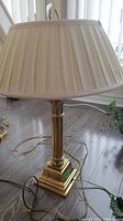 Full view of the brass table lamp with cream fabric shade, showing the lamp assembly, cord, and pleated lampshade with visible wear.