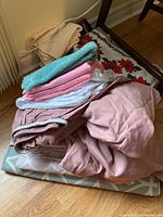Stack of assorted linens including beige fitted sheet, pink sheets, mint green towel, white sheet with zipper, and blue patterned fabric under them.