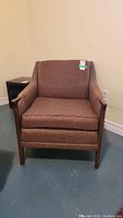 Front view of a mid century modern arm chair with brown upholstery and wooden arms and legs on a blue floor next to a beige wall.