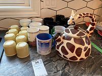 Wide shot showing the ceramic giraffe patterned teapot and multiple mugs of various styles and colors on a countertop.