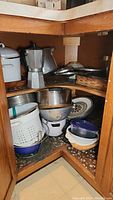 Corner cabinet with stacked cookware including colanders, metal pots, mixing bowls, enamel pots, espresso maker, and baking sheets.
