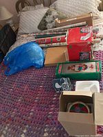 Photo showing multiple Christmas decoration boxes, wrapping paper rolls, and a blue plastic bag on a bed with a purple crocheted throw.