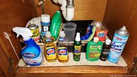 Overview photo showing multiple bottles and cleaning items including Lysol disinfectant spray, Clorox cleaner spray, two bottles of Ajax dish soap, Noxon metal polish, Thom McAn leather conditioner, Brillo pads, cellulose sponge cloths, fruit & vegetable wash, Tuff Stuff spray, SD-20 wash cloths, and a dish tub.