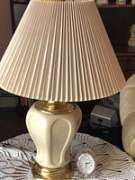 View of cream and gold table lamp with large pleated beige lampshade alongside crystal quartz clock on a lace doily-covered table.