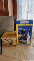 Photo showing original boxes for Marcato Atlas pasta maker and J.A. Henckels Everedge Plus cutlery set with block on marbled countertop near window.
