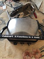 Photo showing Cuisinart Griddle Gourmet stainless steel cooking appliance with logo and 5 functions in 1 unit labeling. Deli slicer visible behind it.