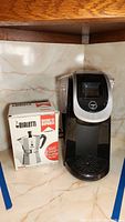 Keurig 2.0 coffee pod machine next to Bialetti Moka Express espresso maker box on a tiled countertop.