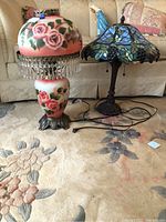 Two decorative lamps side by side on a floral rug, one with pink rose painted glass and crystal fringe, the other with a stained glass shade with grape clusters