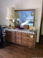 Wooden dresser with nine drawers, dark wood finish, topped with a lace doily and decorative lamp and items, attached mirror reflecting bedroom.