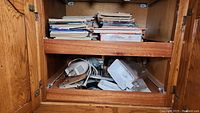 Inside wood cabinet showing stacked magazines, cookbooks, and some kitchen appliances. Items appear mingled and some packing material visible.