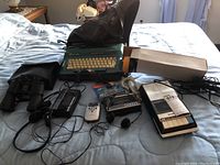 Overview of lot on bed showing all items including typewriter, binoculars, cassette players, remote and cables