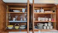 Wood cabinet shelves containing assorted stoneware mugs, plates, bowls, teacups, stemware, basket and other kitchenware