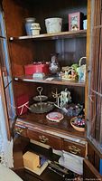 Wood cabinet shelf displaying various ceramic candle holders, floral pot, small holiday figurines, two-tier metal stand, and books underneath.