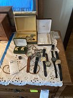 Full view of lot items on a white embroidered cloth including six men's wristwatches, sterling silver ring, vintage coins, cufflinks or earrings, and pen in box.