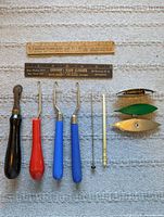 Photo shows antique craft tools and knitting implements arranged on fabric background: 1 dark wood-handled tracing wheel, 3 latch hook tools with black, red, and blue wooden handles, 3 tatting shuttles (two silver, one green), two vintage cardboard rulers, a hat pin, and a small bone or ivory knitting needle.