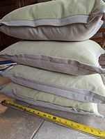 Stack of four cushions showing thickness and two-tone linen-type fabric in pale yellow and taupe with beige piping edges. Measuring tape indicates approx. 20 inches width.