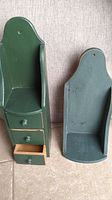 Two wooden wall-mounted shelves, one dark green with three open mini drawers, the other plain and distressed painted greenish-blue, both have a curved top and holes for hanging on wall.
