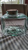 Square glass canister with cork lid sitting on striped fabric, side view showing the canister's shape and cork lid.