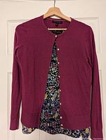 Purple button-up cardigan over floral patterned tank top hanging on a wood hanger against a white door.