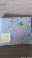 Front of the scrapbook showing blue cover with colorful butterfly illustrations and a sticky note indicating measurements.
