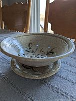 Side angled view of footed pottery fruit bowl with painted decoration and drainage holes, sitting on matching footed plate.