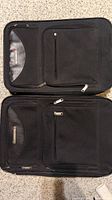 Two black soft fabric carry-on luggage pieces side by side on carpet floor showing front zippered pockets and brand label.